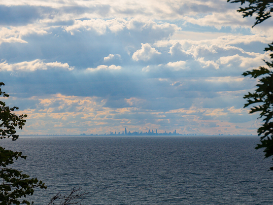 Looking back at the Chicago skyline from afar, through much of Lake Michigan. It was cloudy and the sun casts rays through the cloud. The scene is flanked by silhouettes of trees.