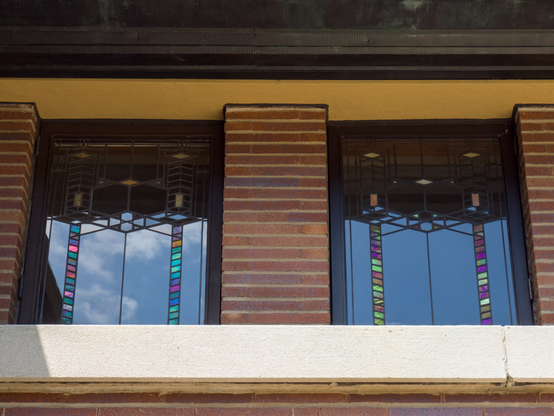 Symmetric composition looking up at two windows of a brick building. The windows are made of geometric pieces of glass, and each of them has two stripes made of colorful glass.