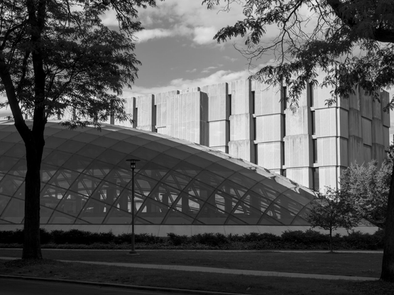 Somewhat abstract black and white photo of a large flat glass dome in front of an old modernist building. The dome reflects the other building.