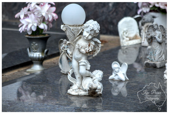 A weathered, white statue of a cherubic angel stands on a dark, reflective granite grave marker, holding a spherical, frosted white glass lamp shade above its head.

A smaller cherub figure rests near its feet. To the left, a silver funerary vase holds a bouquet of white and pink flowers. Other small memorial statues and headstones are softly visible in the background.