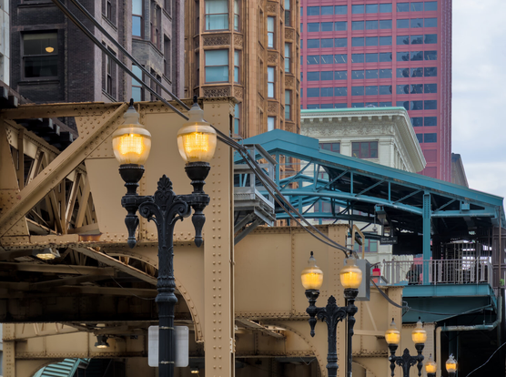 Old style street lights in front of the steel structure of the elevated L train that is painted yellow. There's a L station nearby, which has turquoise metal structure. Behind the station are some early 20th century mid-rise apartment buildings. It was cloudy.
