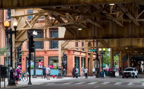 Beneath the L structure that is painted yellow, looking at the sidewalk at an intersection. There're some people on the side walk. There's a building that is painted orange behind them.