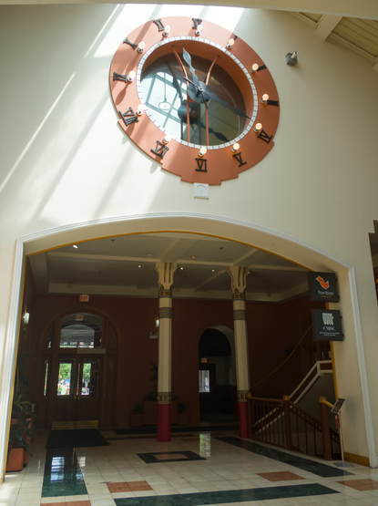 The sun shines through the glass roof on a glass clock in a white wall above the entrance and staircase of the building.