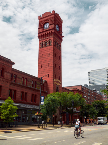 A person is riding a bike on the street beneath the tall clock tower of Dearborn Station. It's a red brick building. Mostly cloudy.