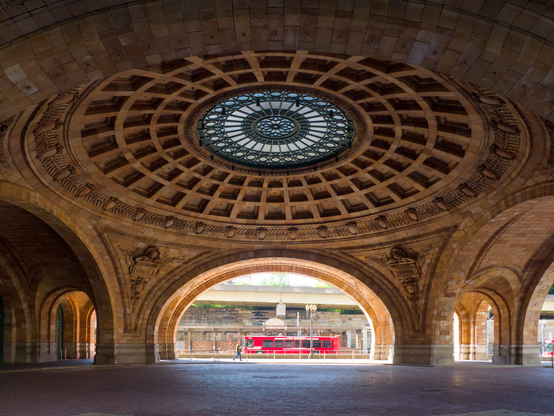 Beneath a great elaborate Beaux Art dome with a glass medallion in the middle. Beneath the arch of the dome in the distance, at least 20 meters away, is a person walking and a red bus.