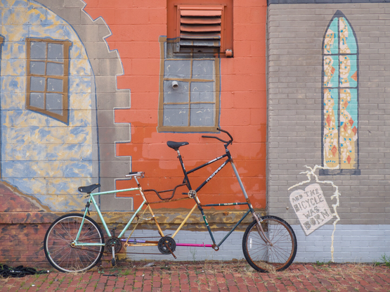 A weird bike that looks like two bikes welded together leans against a wall that has a mural depicting the interior of a castle. The mural features a tablet that says, "And the bicycle was born."