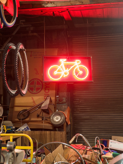 A red neon sign depicting a road bike, above a pile of scrap metal and cardboard and next to some bike tires hung on the wall.