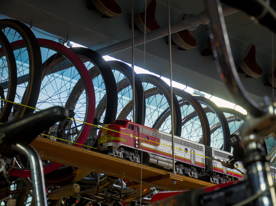 A model of a Santa Fe F7 locomotive pulls a train on a wooden track hung above, passing by many bike tires. The model train is framed by a bike in the foreground, which is out of focus.