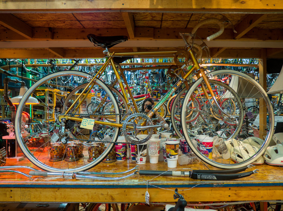 A yellow vintage steel road bike sits by a mirror, which reflects me taking this photo and the colorful pile of bikes and bike parts behind me.