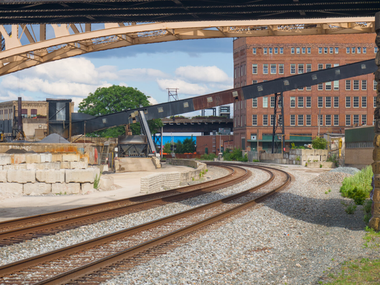 Two railroad tracks curve beneath a yellow steel bridge and a inclined conveyor belt. There's a big red brick building in the background.