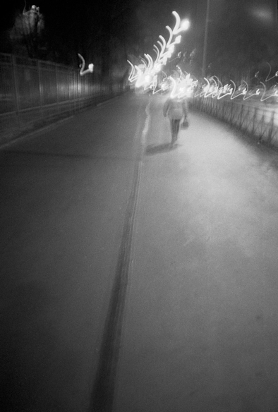 The black-and-white image shows a road that a person is walking along (taken with a long exposure in motion). The lights of the lanterns "blur", the road and the person double - this gives the composition mobility and ghostliness.