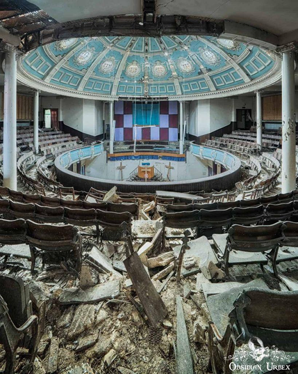 wide view of a circular, amphitheater-style auditorium in a state of decay.
Key Features:
A prominent, ornate, light-blue dome ceiling with decorative plasterwork is visible at the top. 
Rows of dilapidated wooden seating encircle a central floor area.