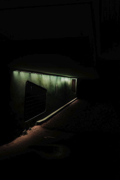 Photo format portrait en couleur, de nuit, on y voit un mur vert avec une grille presque carré, le bord haut descendant de gauche à droite, éclairé par des LED blafardes