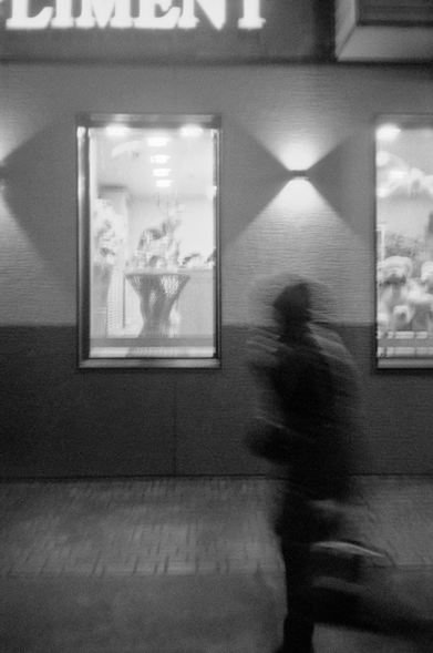 In the black-and-white image, a dark, blurred figure of a man walks against the background of glowing windows and lanterns on the walls.