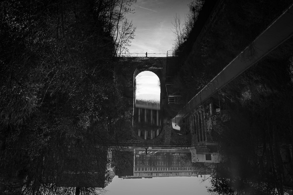 The image is a black and white photograph depicting a bridge over a body of water, with a person standing on top of it. The composition is striking, as the photograph is rotated, giving the impression of looking at a reflection or a distorted perspective.

The archway in the center of the image is the focal point. Through the arch, there is a view of what could be buildings or a cityscape, but it is somewhat blurred. The stonework of the arch is visible, and there are some elements on the side of the arch, perhaps stairs or other structures. A person, appearing as a small silhouette, is standing on the top of the archway or bridge, adding a sense of scale and human presence.
