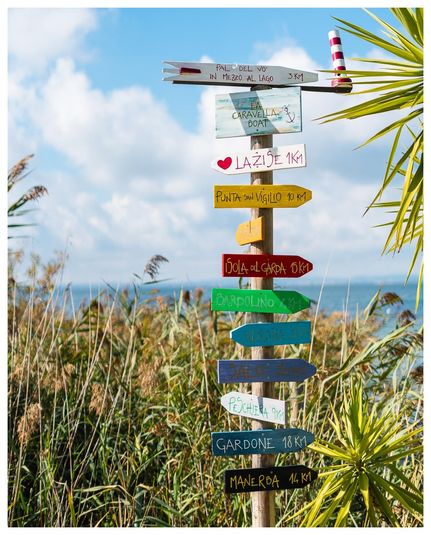 Ein Wegweiser mit kleinen bunten Schildern auf denen Richtung und Entfernung zu einigen italienischen Städtchen am Gardasee stehen. Im Hintergrund eine Palme, Schilf und der See.