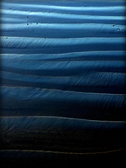 A colour photograph showing a section of sandy beach with light horizontal ripples running through it, rather like the folds of a section of fabric. The sand is a turquoise colour from the blue sky above, graduating from light to dark through the frame.