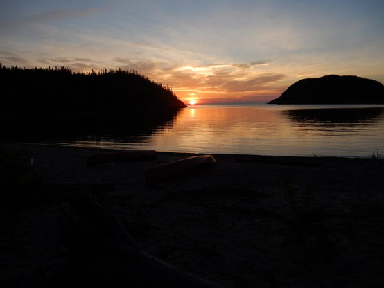 A golden sunset over a large body of water with a hilly peninsula covered in spruce trees on the left, and a large high elevation island on the right. A beach with a canoe on it is in the foreground. Everything is in dark shadow.