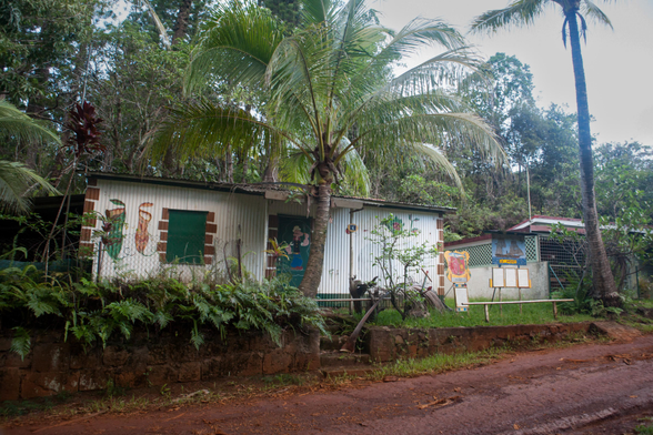 A rustic building surrounded by lush vegetation, featuring palm trees and ferns. The exterior has colorful murals, and there are informational boards nearby, with a painting of the Prony Corvette.