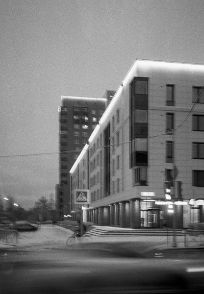 The black-and-white image shows an urban landscape with tall buildings and a road in front of them. Blurred cars race along the road. The blurred figure of a cyclist is also visible.