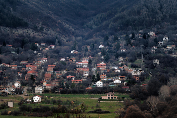 Photo of a distant Balkan mountain village, with a green field in the foreground and a gloomy dark mountain in the background.