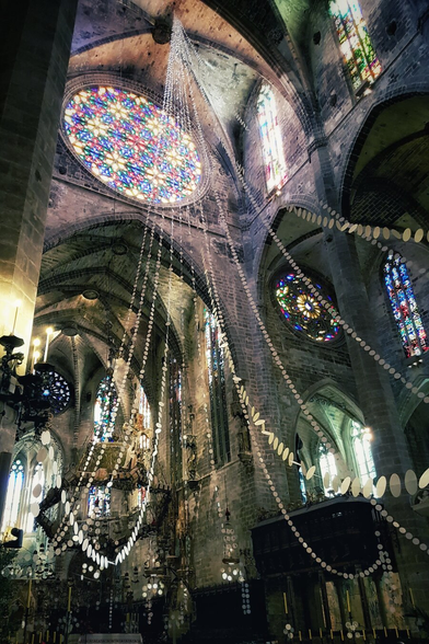 A low-angle interior shot of the Cathedral of Santa Maria of Palma, showcasing towering stone arches and a vaulted ceiling. A magnificent rose window made of vibrant stained glass in hues of red, yellow, and blue illuminates the upper left, casting a soft glow on the masonry. Vertical stained glass windows line the walls on the right. Suspended from the ceiling is a large, artistic installation featuring strings of small, flat discs that drape downwards, adding a modern texture to the ancient Gothic architecture.