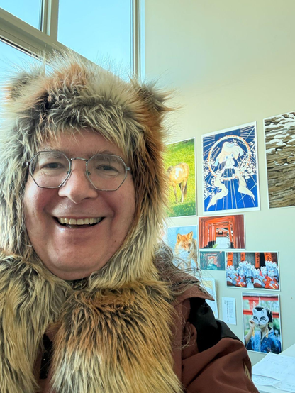 Smiling man with a red fox hood with small ears and a russet color coat is in a corner with windows showing blue sky on the left and prints of: a horse, a Pokemon character, a stone egg, a red fox in a snow and several images of an Inari shrine including a woman in a kimono wearing a white fox mask
