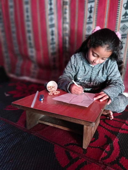 A young girl (Aylan), wearing a gray hooded sweatshirt, sits on the floor drawing a shape on a piece of paper resting on a small wooden table. Pink floral hair clips hold back her long, dark hair. A cup with nuts, a glue stick, and a stapler are on the table. The background features a patterned red and white curtain and a red and black patterned rug.