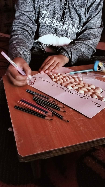 Aylan is drawing with a pink pen on a pink paper, decorated with peanuts, and writing in cursive. Multiple pencils are placed on the surface.
