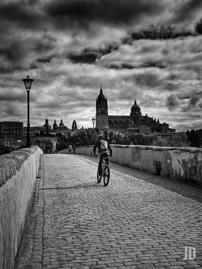 Fotografía en blanco y negro que presenta una perspectiva lineal que guía la mirada hacia un complejo arquitectónico histórico.
Un puente de adoquines (que parece ser el Puente Romano de Salamanca) cruza la imagen desde el primer plano hacia el fondo.
Un ciclista recorre el puente de espaldas, aportando una sensación de movimiento y escala a la escena.
Al fondo se impone la silueta de una catedral con sus cúpulas y torres detalladas contra un cielo muy contrastado.
La iluminación y el procesamiento de la imagen enfatizan el relieve de las piedras y el dramatismo de la formación nubosa.