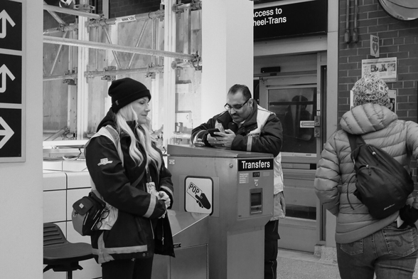 Street level at the station. There is construction in the background. At left, a woman in TTC uniform stands, hands in pockets, a pensive look on her face. Just right of centre, a man in the same uniform leans over his phone, intent. At right, we see the back of someone in winter coat, back to the camera.