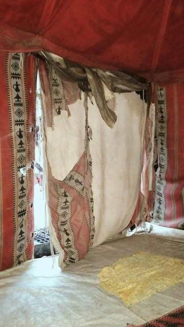 Interior view of a traditional tent with red and patterned fabric curtains and torn, light-colored panels revealing the tent's structure, a light-colored ground covering, and a piece of flat, yellowish carpet.