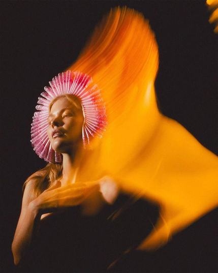 A person with a colorful, ruffled headpiece poses dramatically against a dark background, surrounded by swirling orange lighting.