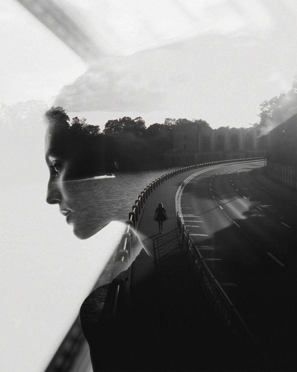 A black and white double-exposure image of a woman framing a shadowy figure walking along a curving pathway beside a body of water, with trees and a road visible in the background.