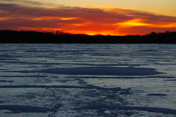 Sunset photograph of orange and yellow clouds over a frozen lake white snow and ice forming white and blue patterns. A face may be visible in the photo from the pattern of clouds and light and snow, if you're prone to that sort of thing.