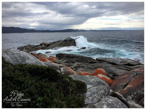 A dramatic, low angle shot of the Binalong Bay coastline under a stormy sky. Large waves are crashing and breaking white against a cluster of dark, rounded granite boulders in the middle ground.

In the foreground, dark green coastal scrub provides a frame, surrounded by rocks stained with bright orange lichen. Distant dark hills line the far horizon.