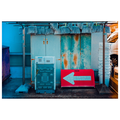 A color photograph of an outdoor utility area. An old washing machine stands on the ground in front of weathered metal doors stained with rust. Above it hang rows of blue mop heads on a metal rack. To the right, a bright red sign with a large white arrow points left, leaning casually against the wall.