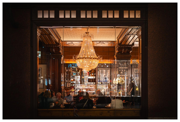 Blick in ein elegantes Café durch ein großes Fenster. Von der aufwändig dekorierten Decke hängt ein überbordender Lüster, der warmes Licht in den Raum  und auf die meist betagten Gäste wirft. In den Fensterscheiben spiegeln sich Schaufensterpuppen und sehen dabei so aus, als stünden sie im Cafe.