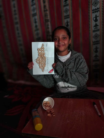 A smiling girl holding a drawing of the map of Palestine, filled with pistachio shells, with the word "Palestine" written vertically alongside the map, sitting on a table with pistachios and art supplies.