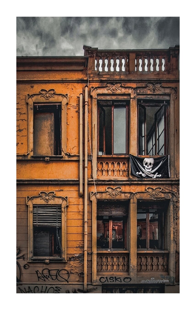 This photo is the facade of an old, orange-toned building with intricate architectural details. It's of the upper two floors and has 6 windows. Some with closed blinds and others open. On one of the balconies on the right, a pirate flag with a skull and crossbones is displayed. The lower part of the building is covered in graffiti. The sky above the building appears overcast.