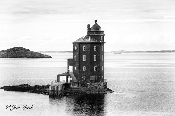 This is a black and white maritime photo of a lighthouse in landscape format. Bjugnfjorden, Norway (2017).

Centred in the image is an octagonal, brick built, lighthouse set on a tiny, rocky  islet in coastal waters. The sea surrounding the lighthouse is dead calm with only the smallest ripples disturbing the almost smooth surface. The sea covers the image from the base to a little above the halfway-up point where instead of a horizon between the sea and sky, there is a long and low level island that stretches from the left to the right margins. Our lighthouse is built on a islet that's about 10 metres long, near tear-drop shaped and rises no more that a metre above the water. This lighthouse is unusual, rather than being tall, imposing and column like - this one is octagonal, brick built and slightly 'house like'. The structure has 4 floors with white framed, rectangular windows on each of the 8 sides on the 3 upper floors, an octagonal pitched, tiled roof and a short lighthouse lantern on the right side. At the base of the lighthouse is a short, 3 or 4 metre, concrete jetty that stretches towards the camera, while on the right side there are two lines running out into the water, perhaps for mooring?

The lighthouse is the iconic Norwegian Kjeungskjær Lighthouse (Kjeungskjær Fyr). Like so many Norwegian lighthouses, it is unlit for part of the summer on account of the midnight sun.