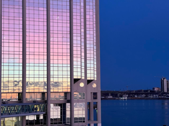 A glass windowed office building reflects a multicoloured sunrise sky. The background harbour and sky are a deep blue with a few buildings dotting the horizon. The colours on the foreground building go from purple at the top through pink, light blue, dark grey, and finally orange. Most windows are rectangular, but two are round. [Halifax (NS, Canada), December 2025]