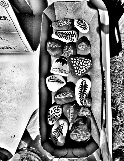 Aus der Vogelperspektive fotografierte Pappschale auf einem Tisch am Straßenrand.
In der Schale liegen kleine bemalte Steine, die mit Punkten, Spiralen oder Federstrichen zumeist abstrakt, dabei vielfältig und sehr harmonisch bemalt sind.