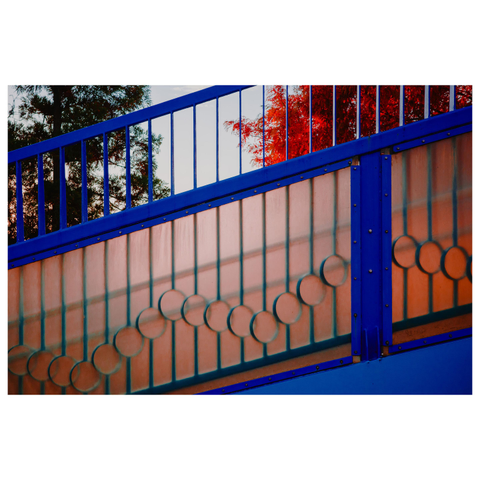 A color photograph of a pedestrian overpass stairway shot from the side. Bright blue railings run diagonally across the frame, with a decorative panel featuring a repeating pattern of circular rings. Behind the railing, trees with vivid red autumn leaves peek through, contrasting with the cool blue metal.