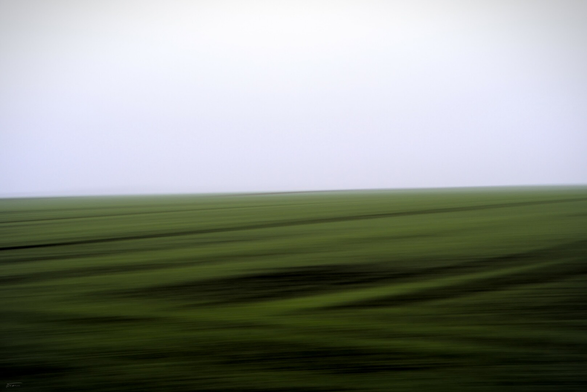 Dieses Foto zeigt eine abstrakte, verschwommene Landschaftsaufnahme, die mit der ICM-Technik (Intentional Camera Movement) erstellt wurde. Die untere Bildhälfte dominieren grüne, schräg verlaufende Streifen, die an ein Feld oder eine Wiese erinnern. Der obere Teil ist hellgrau bis weißlich und wirkt wie Nebel. Die Bewegung der Kamera während der Belichtung erzeugt dynamische, fließende Linien und eine fast malerische Wirkung.