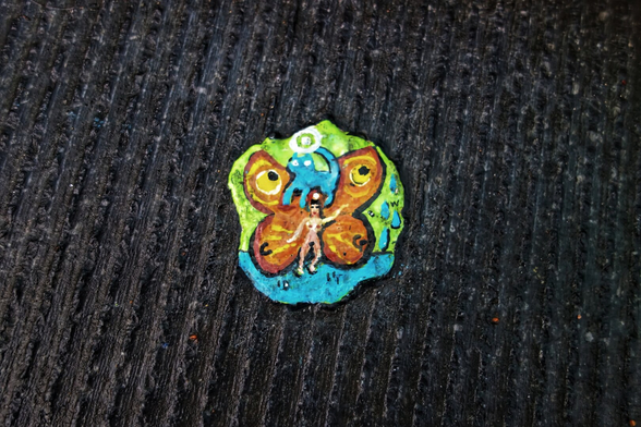 This image features a miniature painting on a piece of discarded chewing gum by British artist Ben Wilson. The artwork depicts a whimsical, surreal scene: a naked woman with butterfly wings, her arms and legs moving as if she is dancing. Her wings are vibrant, with bold orange and yellow patterns outlined in black, and her body is set against a green backdrop and a blue floor.

Above her head floats a blue cat, almost as if it is a halo or a celestial companion. The painting is rich in detail and colour, transforming an ordinary piece of litter into a tiny, imaginative artwork on the pavement.