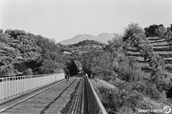 A cyclist rides along the Víaducto Sobre El Arroyo del Higueral surrounded by lush green hills and trees, with distant mountains visible in the background. The image captures a peaceful rural landscape in black and white, emphasizing the natural beauty.
