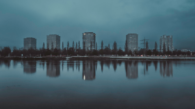 Eine Skyline mit 5 Hochhäuser die sich im nahe gelegenem See auf der Oberfläche spiegeln. Das Ganze ist in einer Art Petrolfarbe eingefärbt.