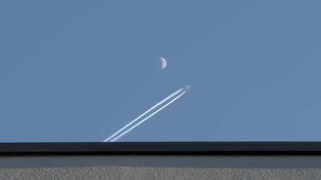 Abstract photo: at the bottom is the edge of a roof, with the sky above it. In the sky, there is a diagonal trace from a plane that is passing the half full moon