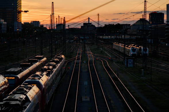 A train station with many tracks, trains on some of them, in front of the setting sun. The trains and the tracks reflect the orange sun light, which is also visible at the top of the image.
